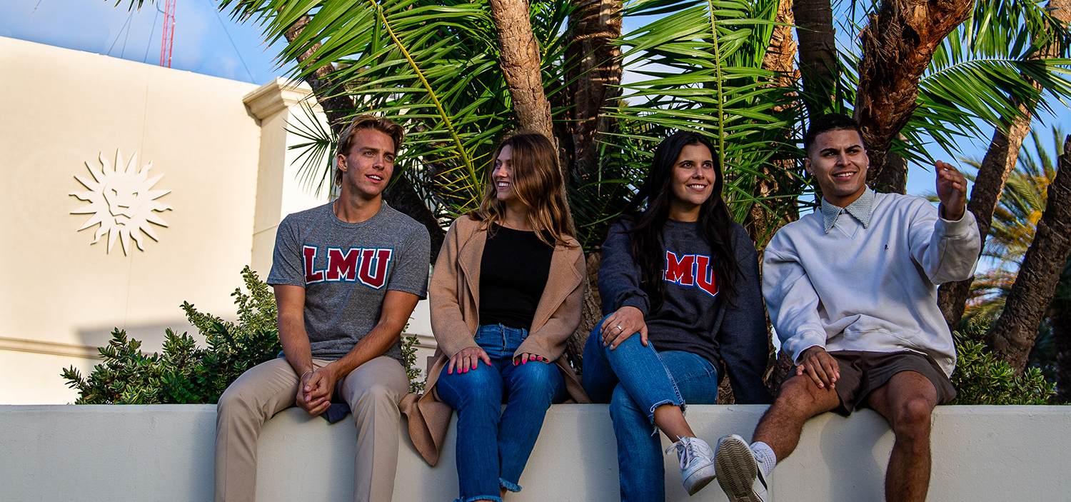 Four students sitting on a low wall, in front of palm trees and a blue sky, two wearing LMU sweatshirts, chatting, with Malone Student Center in the background.